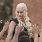 Daenerys Targaryen, com roupa azul e cabelo solto, cercada por seguidores querendo tocar nela, em "Game of Thrones".