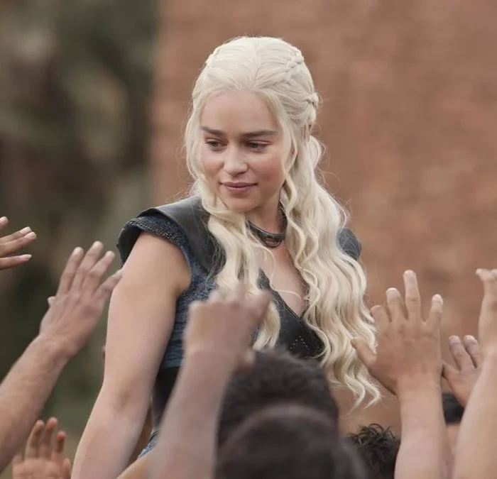 Daenerys Targaryen, com roupa azul e cabelo solto, cercada por seguidores querendo tocar nela, em "Game of Thrones".