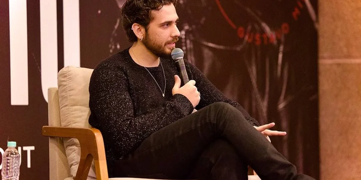 Gustavo Mioto sentado em uma poltrona branca, laterais em madeira, segurando um microfone e olhando para a plateia. Ele está em coletiva de imprensa sobre seu álbum "Atemporal" e há um pôster grande na parede atrás.