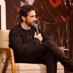 Gustavo Mioto sentado em uma poltrona branca, laterais em madeira, segurando um microfone e olhando para a plateia. Ele está em coletiva de imprensa sobre seu álbum "Atemporal" e há um pôster grande na parede atrás.