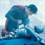 David Corenswet como o homem-de-aço, uniformizado, em "Superman" (2025), de James Gun. Ele está abaixado, agachado com pressão sobre um de seus joelhos, e olhando e lamentando a morte de um robô.