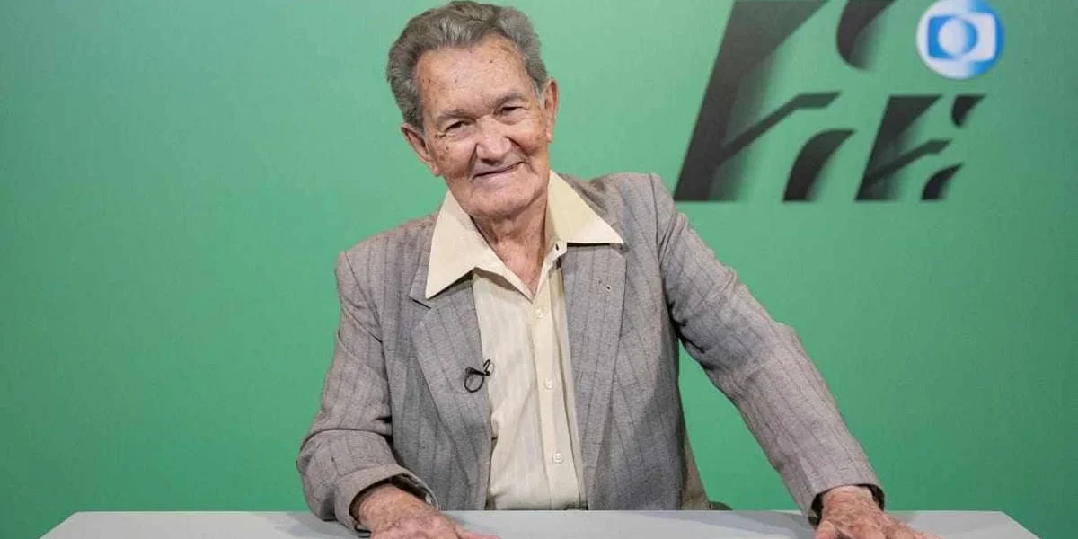 Léo Batista de terno cinza e roupa social creme com o corpo inclinado à sua direita (nossa esquerda), e mãos sobre uma bancada de tom cinza mais claro que sua roupa. Narrador está sorrindo.