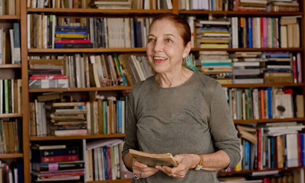 Autora Marina Colasanti sorrindo, de vestido cinza, segurando um livro com as duas mãos e olhando para o interlocutor. Ela está em uma biblioteca.