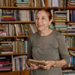 Autora Marina Colasanti sorrindo, de vestido cinza, segurando um livro com as duas mãos e olhando para o interlocutor. Ela está em uma biblioteca.