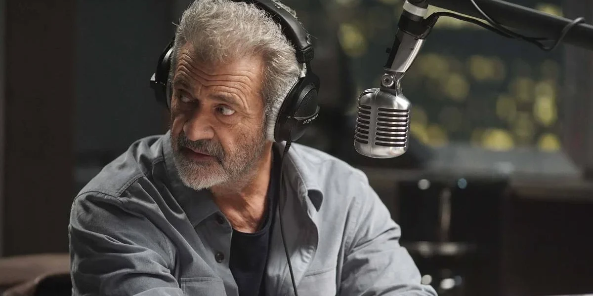 Mel Gibson em "On The Line" ou "Até o Limite" (2022), interpretando o protagonista radialista Elvis Cooney. Ele está na frente de um microfone, virando o roto para sua direita, como se ouvisse ou visse algo atrás de si.