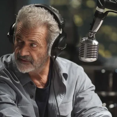 Mel Gibson em "On The Line" ou "Até o Limite" (2022), interpretando o protagonista radialista Elvis Cooney. Ele está na frente de um microfone, virando o roto para sua direita, como se ouvisse ou visse algo atrás de si.
