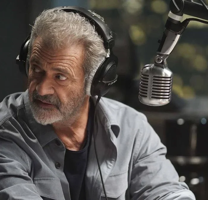 Mel Gibson em "On The Line" ou "Até o Limite" (2022), interpretando o protagonista radialista Elvis Cooney. Ele está na frente de um microfone, virando o roto para sua direita, como se ouvisse ou visse algo atrás de si.