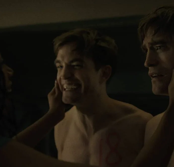 Robert Pattinso como o protagonista de "Mickey 17". Ele está sem camisa nas duas versões, a esquerda, o número 18, sorri de forma estranha e quase desesperada, a da direita, o Mickey 17, está assustada. Naomi Ackie (Nasha Adjaya) aberta a bochecha dos dois.