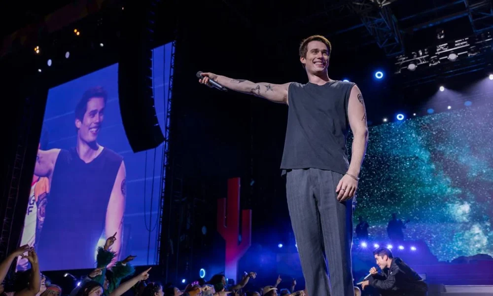 Nicholas Galitzine como o protagonista de "Uma Ideia de Você" (2024), como um cantor no palco com o braço direito estendido e microfone na mão.