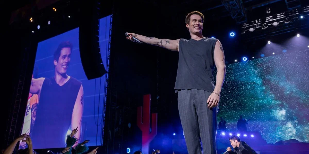 Nicholas Galitzine como o protagonista de "Uma Ideia de Você" (2024), como um cantor no palco com o braço direito estendido e microfone na mão.