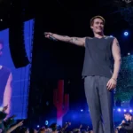 Nicholas Galitzine como o protagonista de "Uma Ideia de Você" (2024), como um cantor no palco com o braço direito estendido e microfone na mão.