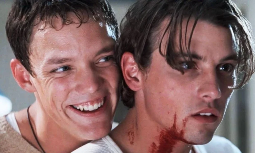 Stu e Billy, os atores Matthew Lillard e Skeet Ulrich, respectivamente à esquerda e direita, no filme "Pânico" (1996). Stu está sorrindo maniacamente sobre o ombro direito de Billy, que está com o pescoço ensanguentado.