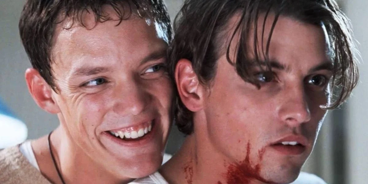 Stu e Billy, os atores Matthew Lillard e Skeet Ulrich, respectivamente à esquerda e direita, no filme "Pânico" (1996). Stu está sorrindo maniacamente sobre o ombro direito de Billy, que está com o pescoço ensanguentado.