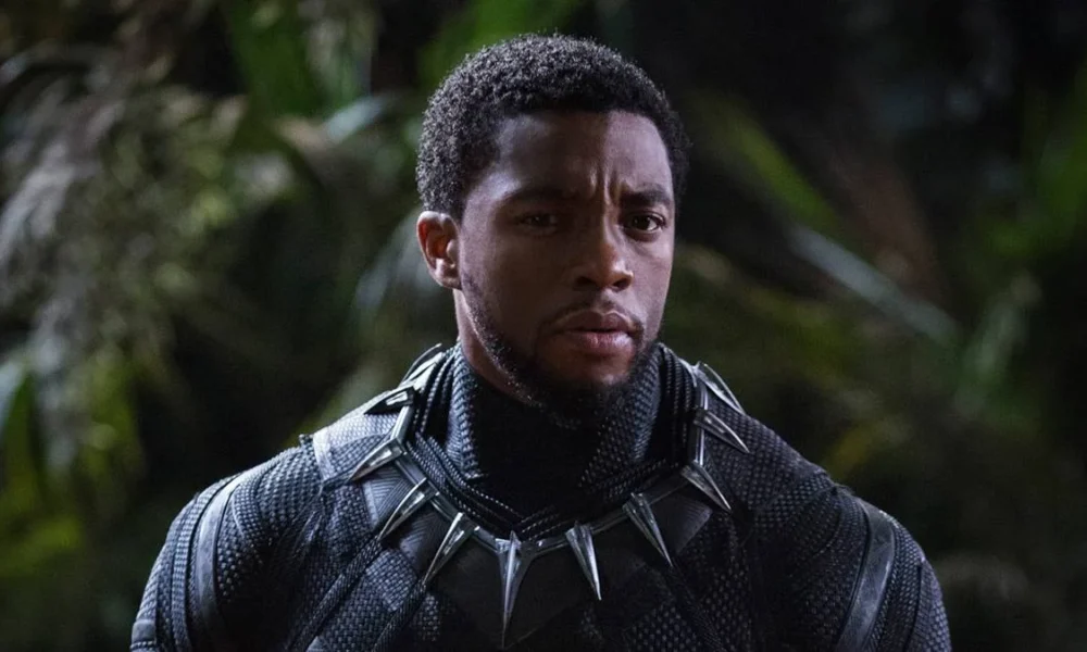 Chadwick Boseman no papel de T'Challa em "Pantera Negra" (2018). Há uma mata desfocada ao fundo e o autor está nos trajes do super-herói, sem apenas a parte da cabeça.
