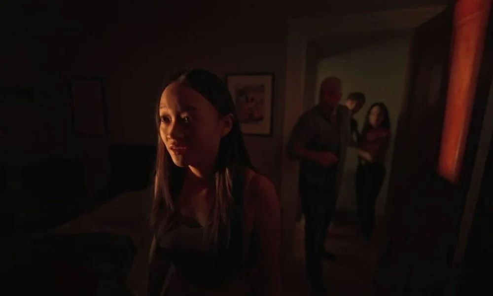Família em casa do terror "Presence" (2025), da Neon. Garota à esquerda está boquiaberta e assustada com algo. Única iluminação do cômodo vem de uma fresta em janela.