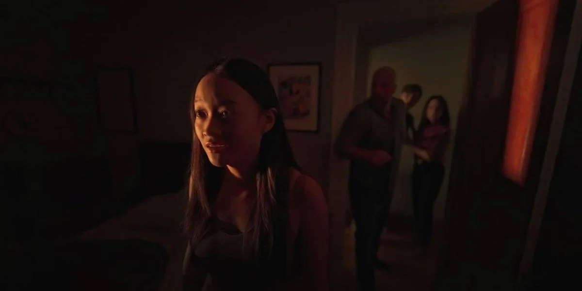 Família em casa do terror "Presence" (2025), da Neon. Garota à esquerda está boquiaberta e assustada com algo. Única iluminação do cômodo vem de uma fresta em janela.
