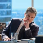 Imagem promocional de "Suits LA" com protagonista com corpo inclinado em mesa de escritório para sua esquerda, apoiando-se no seu braço direito, de terno, e com braço direito sobre a mesa, pensativo.