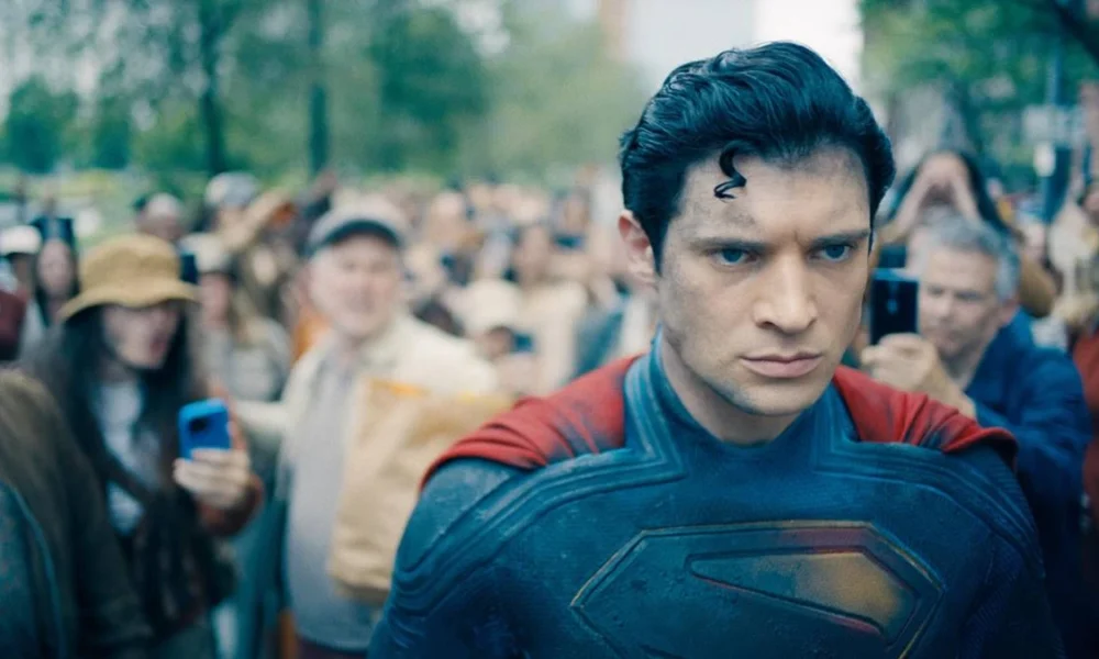 David Corenswet como o protagonista do longa "Superman", de 2025. Personagem está com o traje e cabelo característicos do homem de aço, com um olhar sério, irritadiço, cheio de foligem, e uma multidão o fotografa.