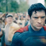 David Corenswet como o protagonista do longa "Superman", de 2025. Personagem está com o traje e cabelo característicos do homem de aço, com um olhar sério, irritadiço, cheio de foligem, e uma multidão o fotografa.