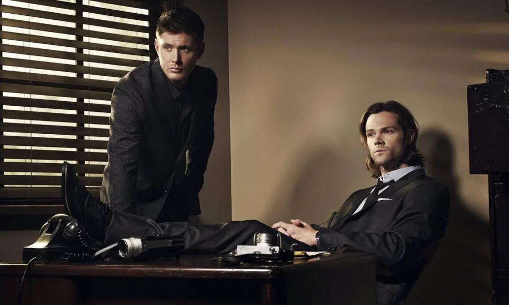 Irmãos Winchester. Dean está apoiado com as palmas da mão em uma escrivaninha, e Sam sentado com as pernas em cima da mesa. Personagens são de "Supernatural".