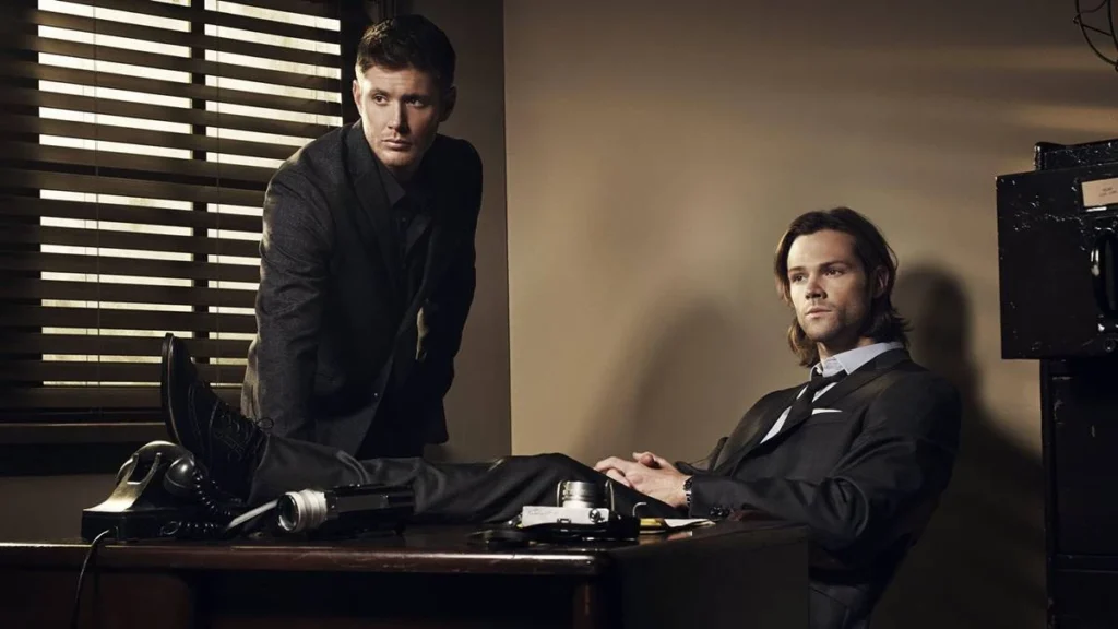 10 Finais Mais Odiados de Todos os Tempos: Do Luxo ao Lixo 1 Irmãos Winchester. Dean está apoiado com as palmas da mão em uma escrivaninha, e Sam sentado com as pernas em cima da mesa. Personagens são de "Supernatural".