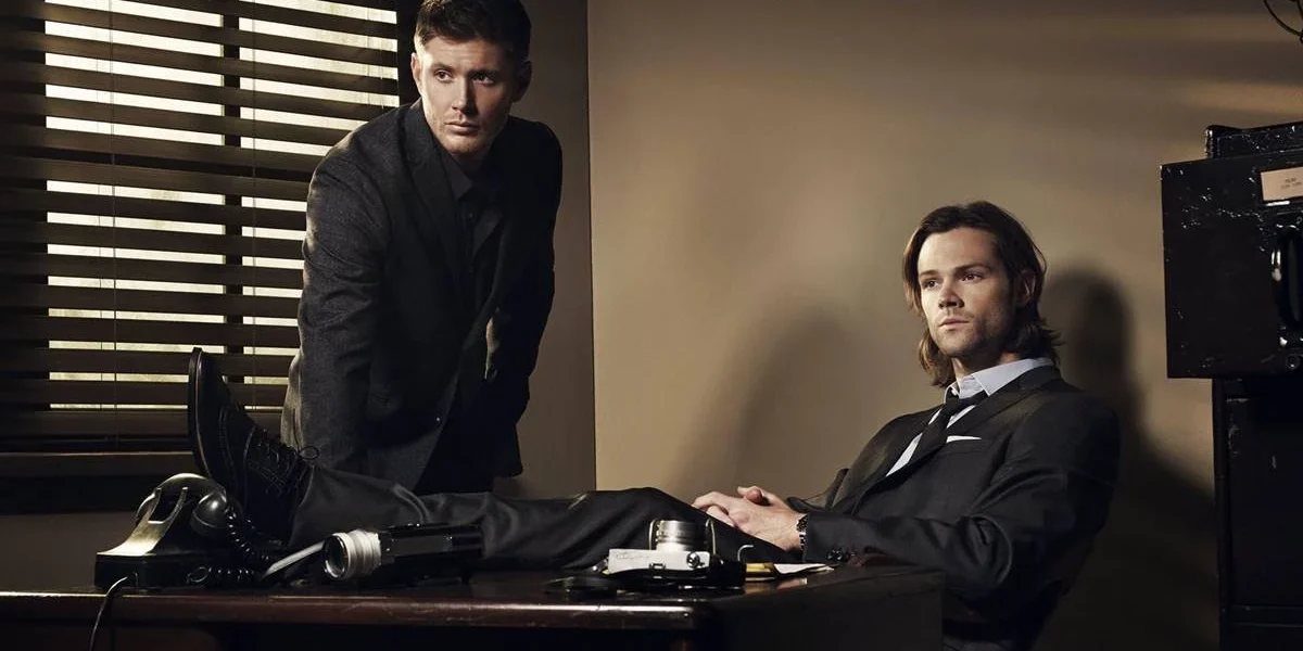 Supernatural | Como “Carry On Wayward Son” se Tornou a Alma da Série Irmãos Winchester. Dean está apoiado com as palmas da mão em uma escrivaninha, e Sam sentado com as pernas em cima da mesa. Personagens são de "Supernatural".