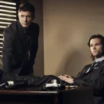 Irmãos Winchester. Dean está apoiado com as palmas da mão em uma escrivaninha, e Sam sentado com as pernas em cima da mesa. Personagens são de "Supernatural".