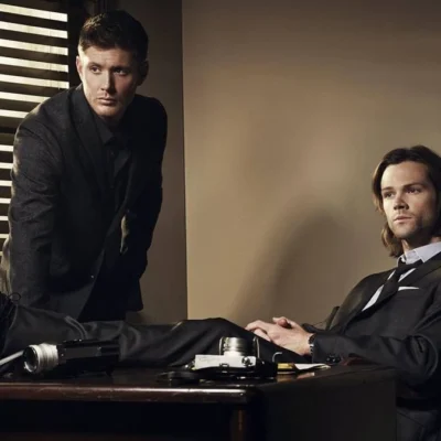 Irmãos Winchester. Dean está apoiado com as palmas da mão em uma escrivaninha, e Sam sentado com as pernas em cima da mesa. Personagens são de "Supernatural".