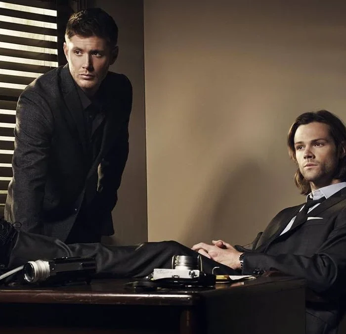 Irmãos Winchester. Dean está apoiado com as palmas da mão em uma escrivaninha, e Sam sentado com as pernas em cima da mesa. Personagens são de "Supernatural".