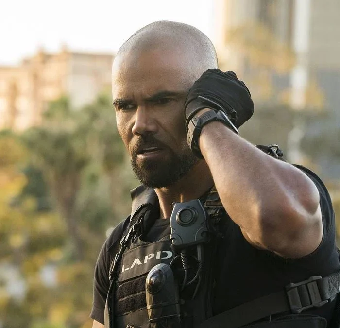 Shemar Moore como Daniel 'Hondo' Harrelson, protagonista de "S.W.A.T.". Fardado, ele coloca a mão na escuta no ouvido esquerdo e tenta prestar atenção. No fundo desfocado, árvores meio a uma cidade.