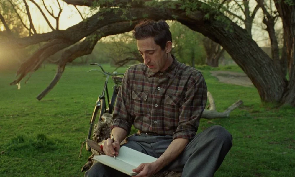 László Tóth (Adrien Brody) sentado debaixo de uma árvore com um caderno em mãos. Imagem do filme "The Brutalist".