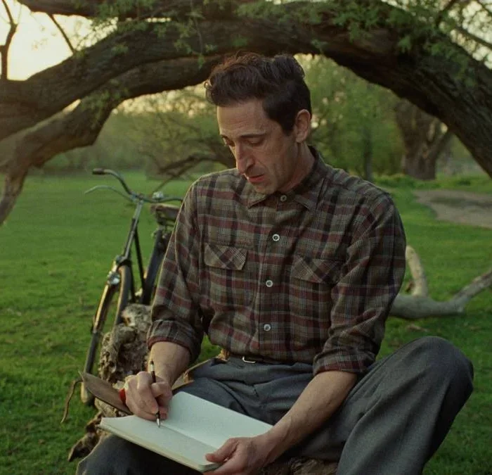 László Tóth (Adrien Brody) sentado debaixo de uma árvore com um caderno em mãos. Imagem do filme "The Brutalist".