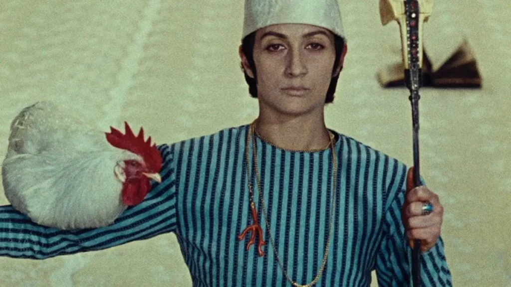 A Cor da Romã (1969) | Se Poesia Fosse Um Filme 1 Um homem com vestes tradicionais armênias em "The Color of Pomegranates" (1969), roupa listrada em dois tons de azul, chapéu branco, e uma galinha no braço direito. Cena de "The Color of Pomegranates" (1969).
