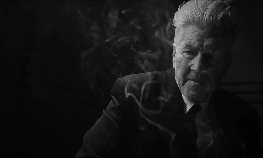 David Lynch em material promocional de "What Did Jack Do?", curta de 2017 em preto e branco. Ele está de terno e podemos ver uma fumaça de cigarro em volta de seu corpo.