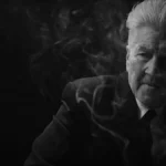 David Lynch em material promocional de "What Did Jack Do?", curta de 2017 em preto e branco. Ele está de terno e podemos ver uma fumaça de cigarro em volta de seu corpo.