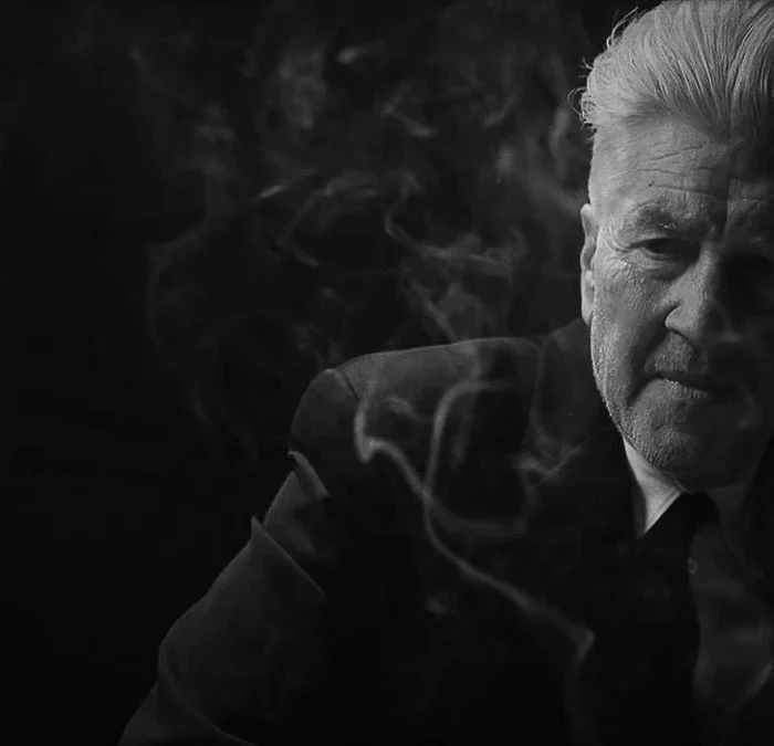 David Lynch em material promocional de "What Did Jack Do?", curta de 2017 em preto e branco. Ele está de terno e podemos ver uma fumaça de cigarro em volta de seu corpo.