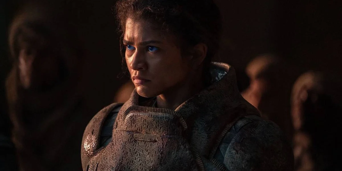 Zendaya como Chani em "Duna: Parte 2" (2024), pela Warner Bros. Personagem está com um semblante sério, olhando fixamente a um ponto. Seus olhos azuis são destaque na imagem dominada por tons escuros.