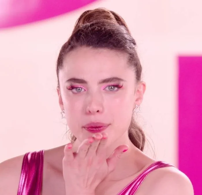 Margaret Qualley como a protagonista de "A Substância" (2024). Ela está com roupas de ginástia rosa, delineado rosa, unhas também rosa, olhando para a câmera e assoprando um beijinho.