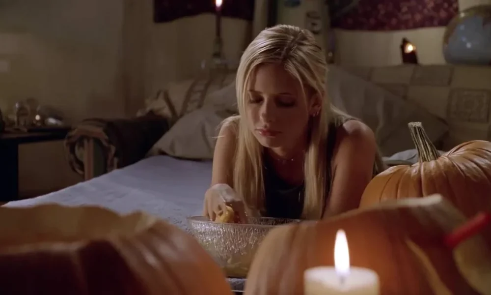 Sarah Michelle Gellar na cama de bruços, apoiada sobre os covotevelos, rodeada de abóboras e com uma bacia na sua frente, a qual ela encara. Atriz está interpretando a protagonista de "Buffy: A Caça Vampiros".