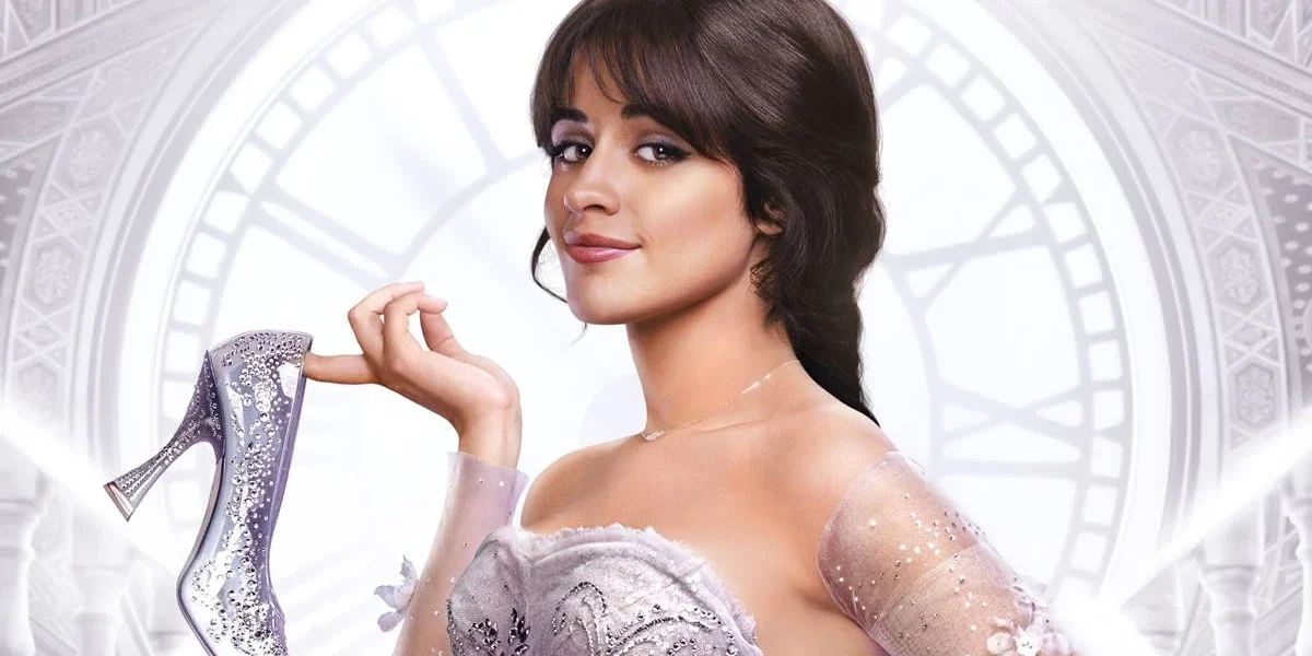 Camila Cabello em imagem promocional como a protagonista de "Cinderella" (2021). Ela segura o sapato de cristal com um dos dedos e sorri para a câmera.