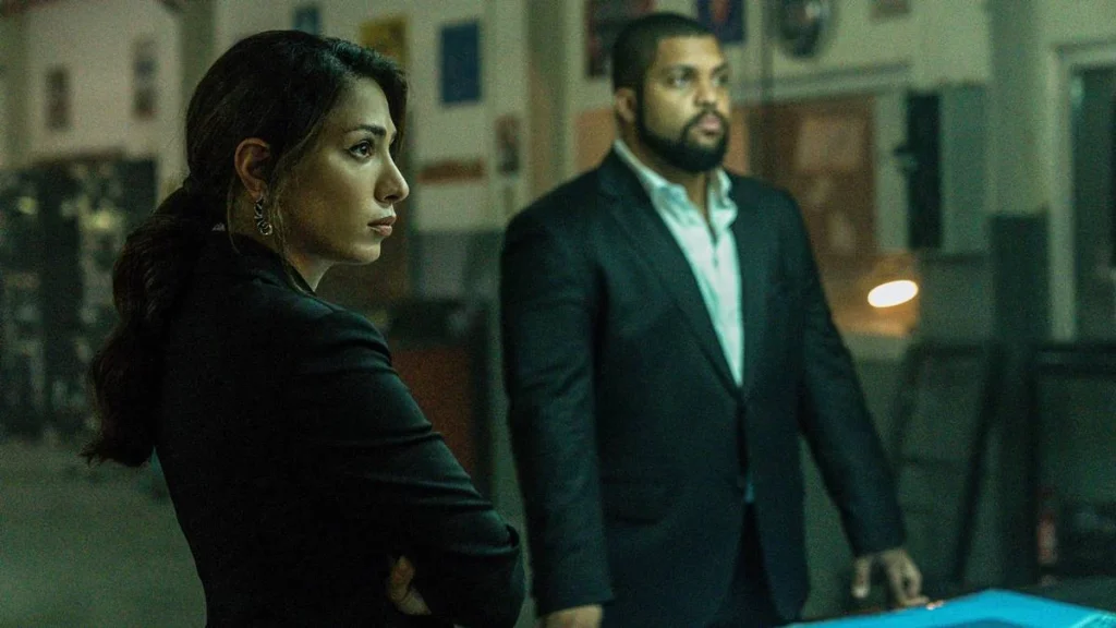 Covil de Ladrões 2 | Tudo Muda Para Continuar o Mesmo 1 Evin Ahmad como Jovanna, à esquerda, e O'Shea Jackson Jr. como Donnie Wilson, à direita, ambos de terno. Ela está de braços cruzados e ambos olham para um mesmo ponto, fora da câmera, à direita. Imagem do filme "Covil de Ladrões 2".