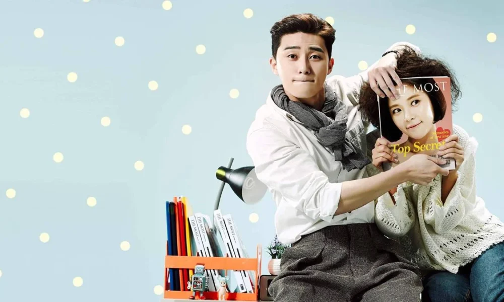 Park Seo-jun (esquerda) e Hwang Jung-eum (direita) para foto promocional de "Ela Era Bonita" ou "She Was Pretty". Ele a ajuda a esconder seu rosto com uma revista, com uma modelo na capa, enquanto apenas seus cabelos estão visíveis quanto ao corpo acima do pescoço. Eles estão sentados em uma escrivaninha.