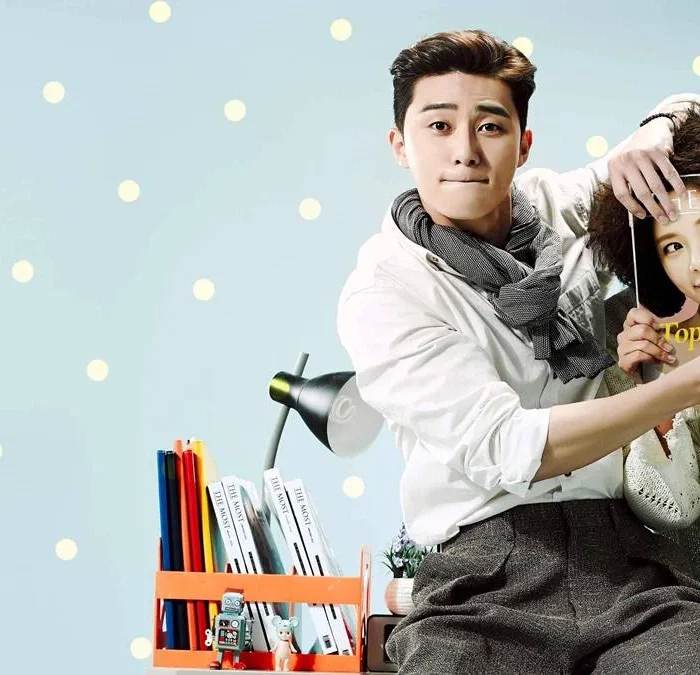 Park Seo-jun (esquerda) e Hwang Jung-eum (direita) para foto promocional de "Ela Era Bonita" ou "She Was Pretty". Ele a ajuda a esconder seu rosto com uma revista, com uma modelo na capa, enquanto apenas seus cabelos estão visíveis quanto ao corpo acima do pescoço. Eles estão sentados em uma escrivaninha.