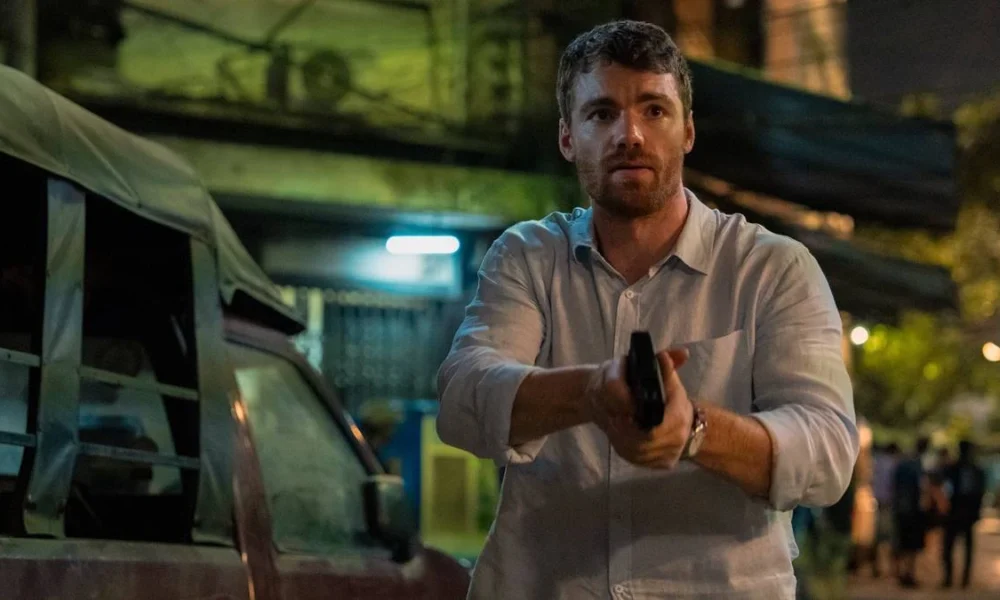 Peter Sutherland (Gabriel Basso) empunhando um revólver com as duas mãos, semblante preocupado, em uma paisagem de subúrbio na segunda temporada de "O Agente Noturno".