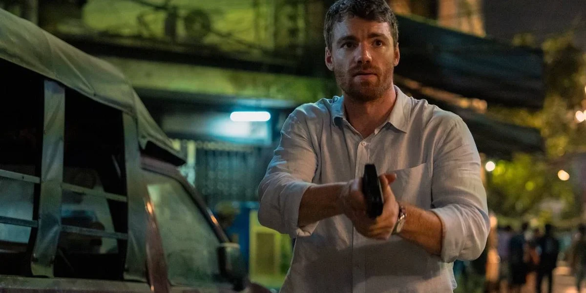 Peter Sutherland (Gabriel Basso) empunhando um revólver com as duas mãos, semblante preocupado, em uma paisagem de subúrbio na segunda temporada de "O Agente Noturno".