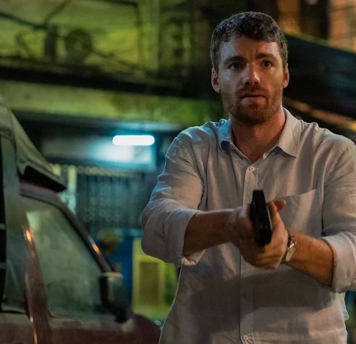 Peter Sutherland (Gabriel Basso) empunhando um revólver com as duas mãos, semblante preocupado, em uma paisagem de subúrbio na segunda temporada de "O Agente Noturno".