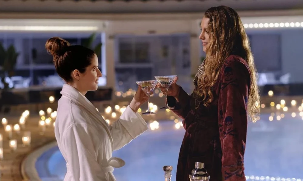 Anna Kendrick e Blake Lively como Stephanie Smothers e Emily Nelson, protagonistas de "Outro Pequeno Favor". Elas estão em se olhando, brindando com as mãos próximas uma da outra, tomando um drink, em um lugar luxuoso desfocado ao fundo.