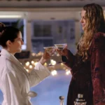 Anna Kendrick e Blake Lively como Stephanie Smothers e Emily Nelson, protagonistas de "Outro Pequeno Favor". Elas estão em se olhando, brindando com as mãos próximas uma da outra, tomando um drink, em um lugar luxuoso desfocado ao fundo.
