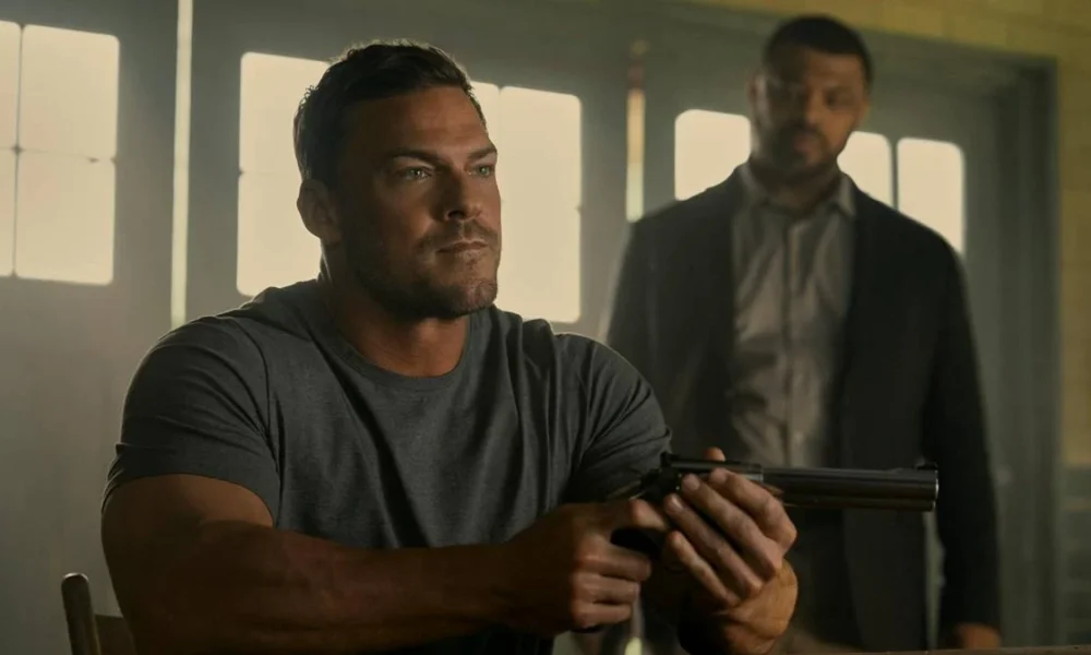 Alan Ritchson como Jack Reacher na terceira temporada de "The Reacher". Ele está sentado em uma mesa, vestido com camiseta lisa cinza e segurando um revólver com as duas mãos enquanto encara alguém do outro lado.