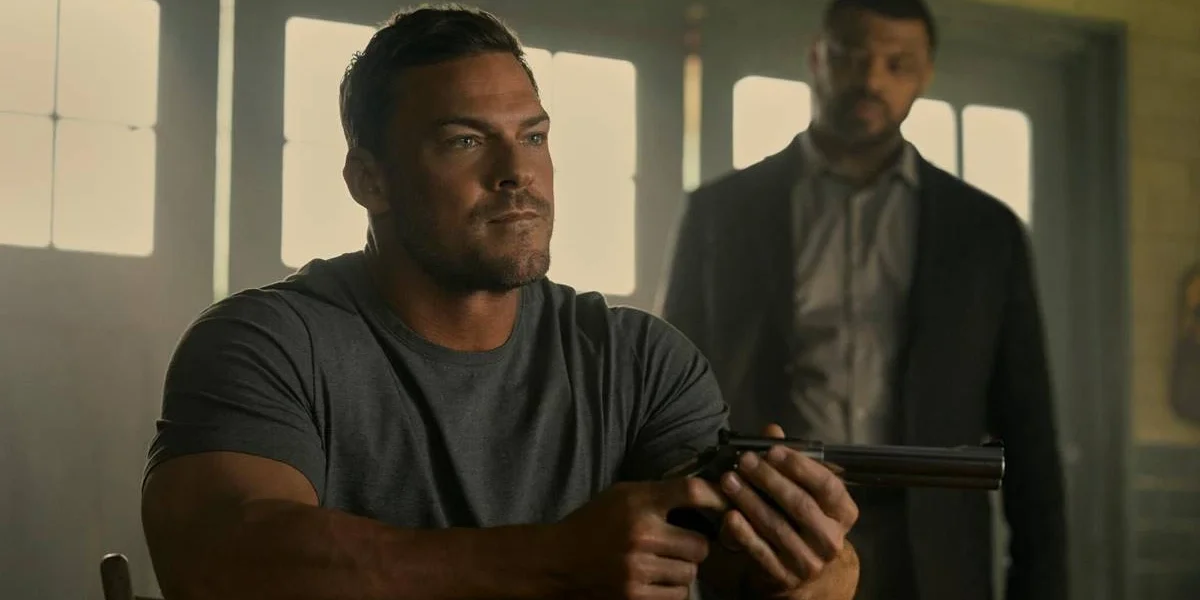 Alan Ritchson como Jack Reacher na terceira temporada de "The Reacher". Ele está sentado em uma mesa, vestido com camiseta lisa cinza e segurando um revólver com as duas mãos enquanto encara alguém do outro lado.